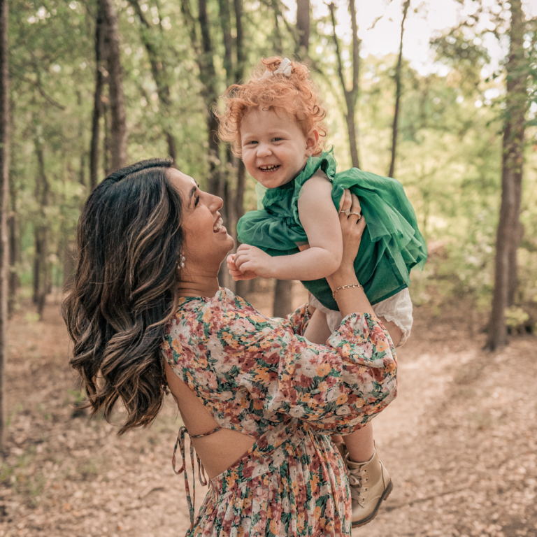 A mother joyfully lifts her smiling toddler in a forest.