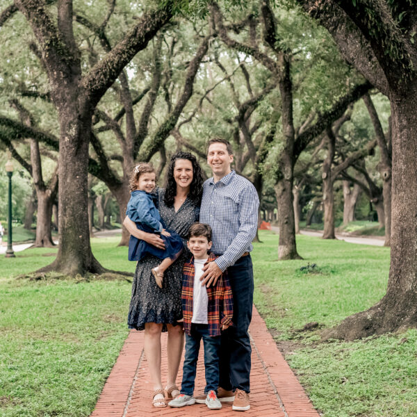 A family of four stands on a tree-lined path, smiling warmly.